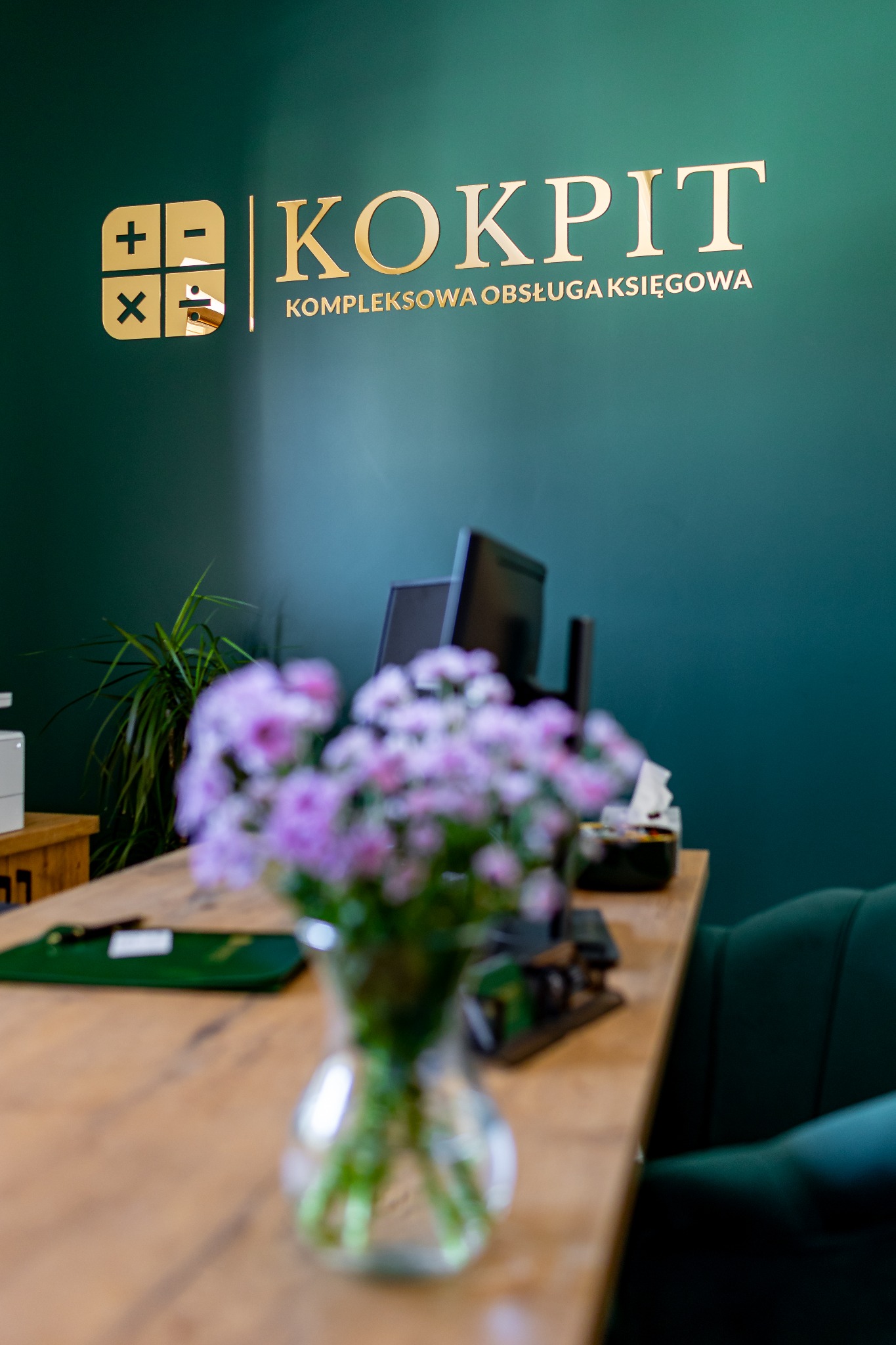 Recepcja biura oferującego kompleksową obsługę finansową, z złotym logo na ciemnozielonej ścianie, bukietem fioletowych kwiatów na drewnianym biurku i widocznymi monitorami w tle.