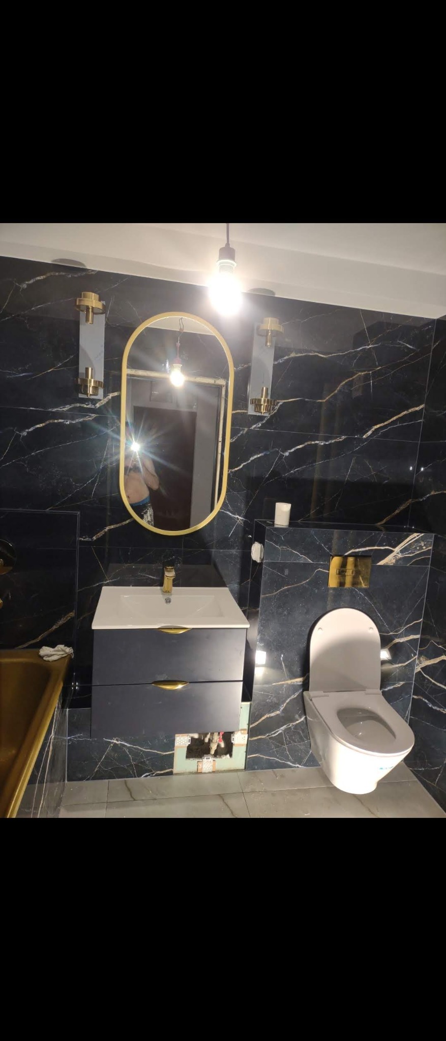 Elegancka łazienka z czarnymi marmurkowymi płytkami, owalnym lustrem w złotej ramie, białą umywalką z szafką w odcieniach szarości i złotymi uchwytami, toaleta w komplecie, oświetlenie kinkietowe...