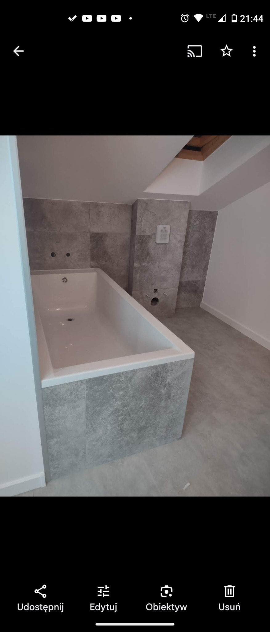 Wanna obudowana szarymi płytkami imitującymi kamień, dopasowanymi do płytek ściennych w łazience na poddaszu ze świetlikiem w suficie.