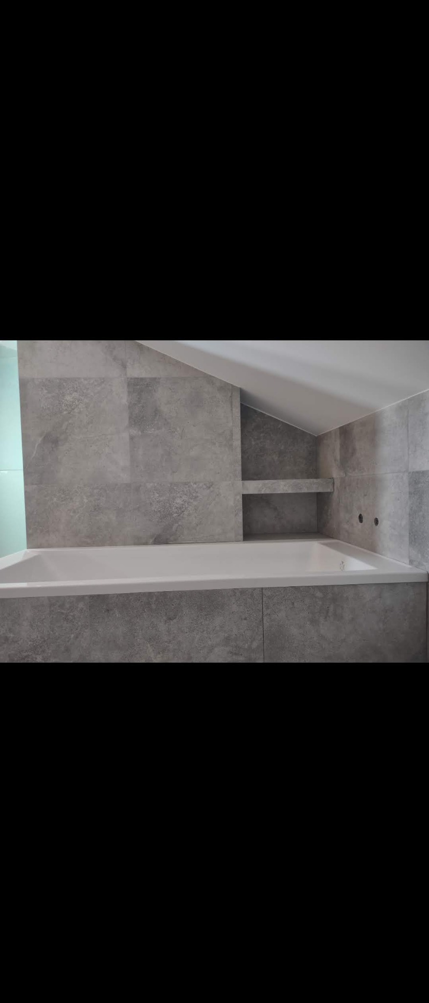 Nowoczesna łazienka w szarościach: wanna obudowana płytkami imitującymi beton, wnęka z półkami pod skosem sufitu, minimalistyczne baterie.