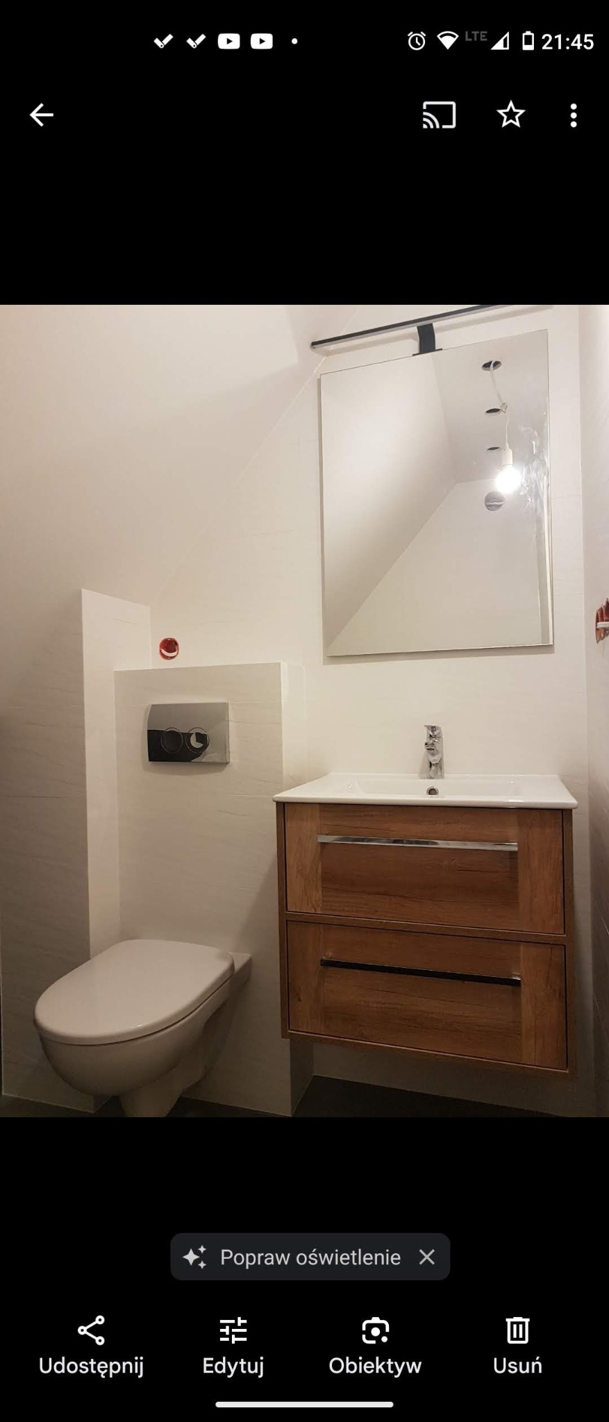 Nowoczesna łazienka z wiszącą toaletą, umywalką z drewnianą szafką i dużym lustrem pod skośnym sufitem, widoczne niedokończone otwory na oświetlenie.