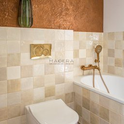 MADERA - Studio Projektowania Wnętrz i dekoracji ścian - Beżowa łazienka z mozaiką, złotą armaturą i dekoracyjną ścianą w odcieniach brązu. Umywalka podwieszana i kwiaty w wazonie dodają elegancji.