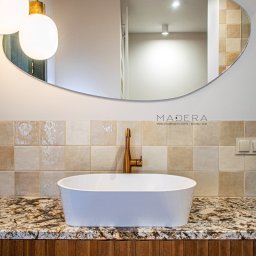 MADERA - Studio Projektowania Wnętrz i dekoracji ścian - Nowoczesna łazienka: biała umywalka na blacie z granitu, złota bateria, lustro o nieregularnym kształcie i beżowe płytki. Oświetlenie punktowe i lampy wiszące.