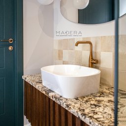 MADERA - Studio Projektowania Wnętrz i dekoracji ścian - Nowoczesna łazienka: biała umywalka na blacie z granitu, złota bateria, lustro i lampy wiszące. Ciemnozielone drzwi dodają elegancji.