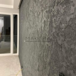 MADERA - Studio Projektowania Wnętrz i dekoracji ścian - Szara ściana z unikalną fakturą, wykończona w stylu dekoracyjnym. Widoczne logo studia projektowania wnętrz 'MADERA' i fragment okna w tle.