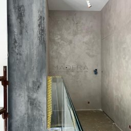 MADERA - Studio Projektowania Wnętrz i dekoracji ścian - Wnętrze z dekoracyjnymi szarymi ścianami wykończonymi w stylu betonu architektonicznego i szklaną balustradą. Widoczne przygotowania do montażu oświetlenia.