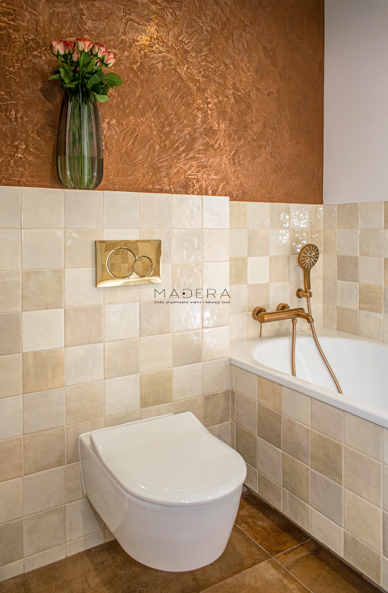 Beżowa łazienka z mozaiką, złotą armaturą i dekoracyjną ścianą w odcieniach brązu. Umywalka podwieszana i kwiaty w wazonie dodają elegancji.