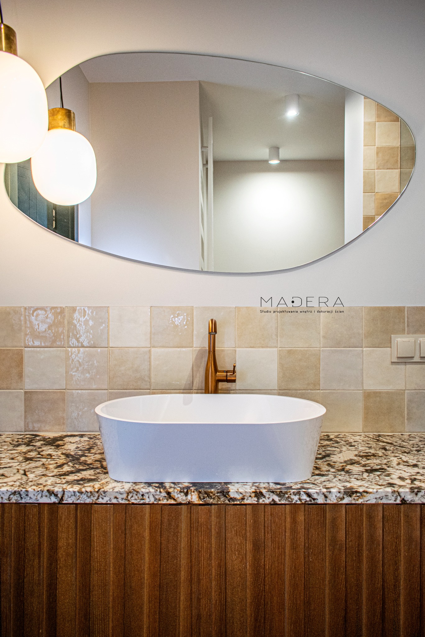 Nowoczesna łazienka: biała umywalka na blacie z granitu, złota bateria, lustro o nieregularnym kształcie i beżowe płytki. Oświetlenie punktowe i lampy wiszące.