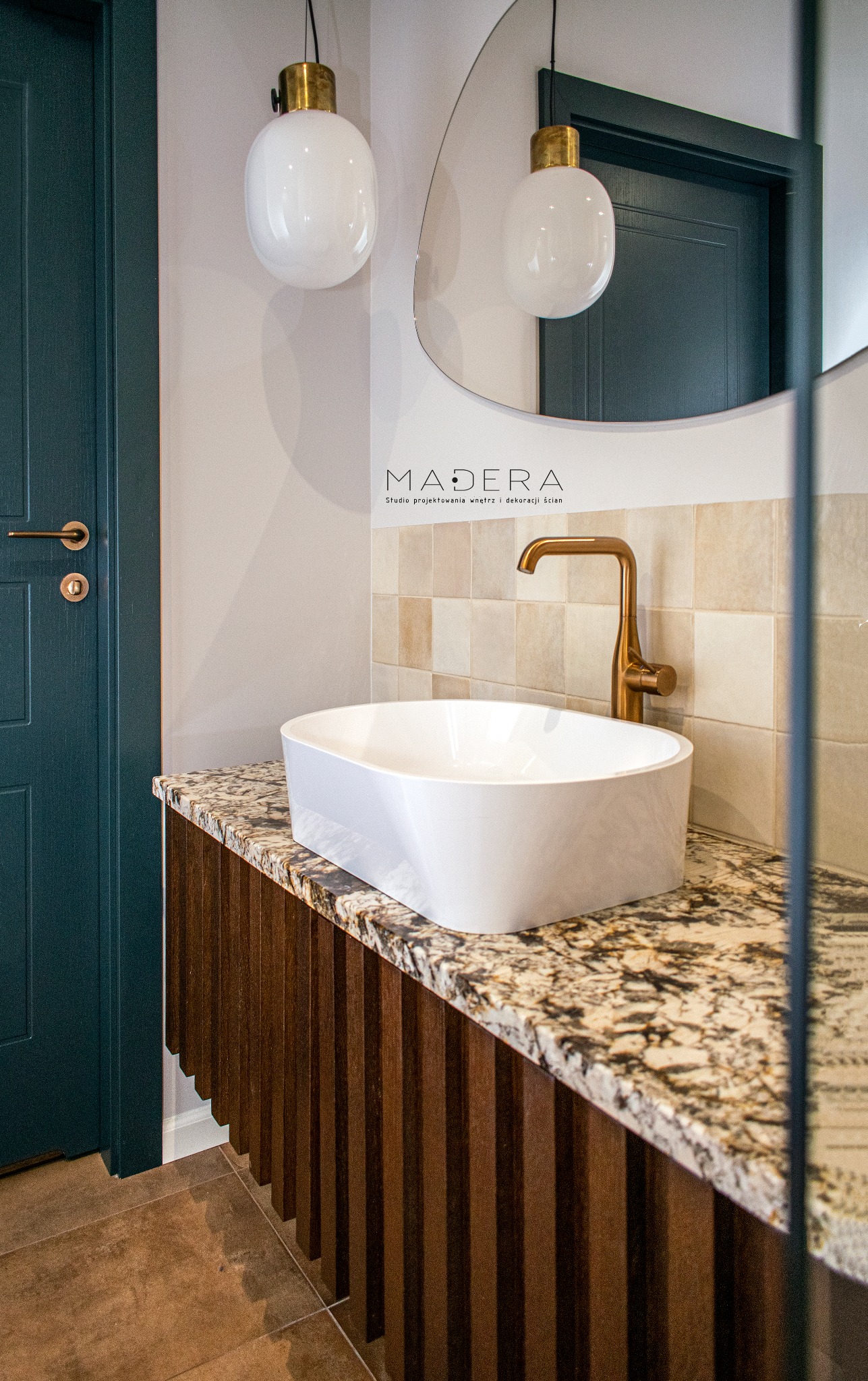 Nowoczesna łazienka: biała umywalka na blacie z granitu, złota bateria, lustro i lampy wiszące. Ciemnozielone drzwi dodają elegancji.