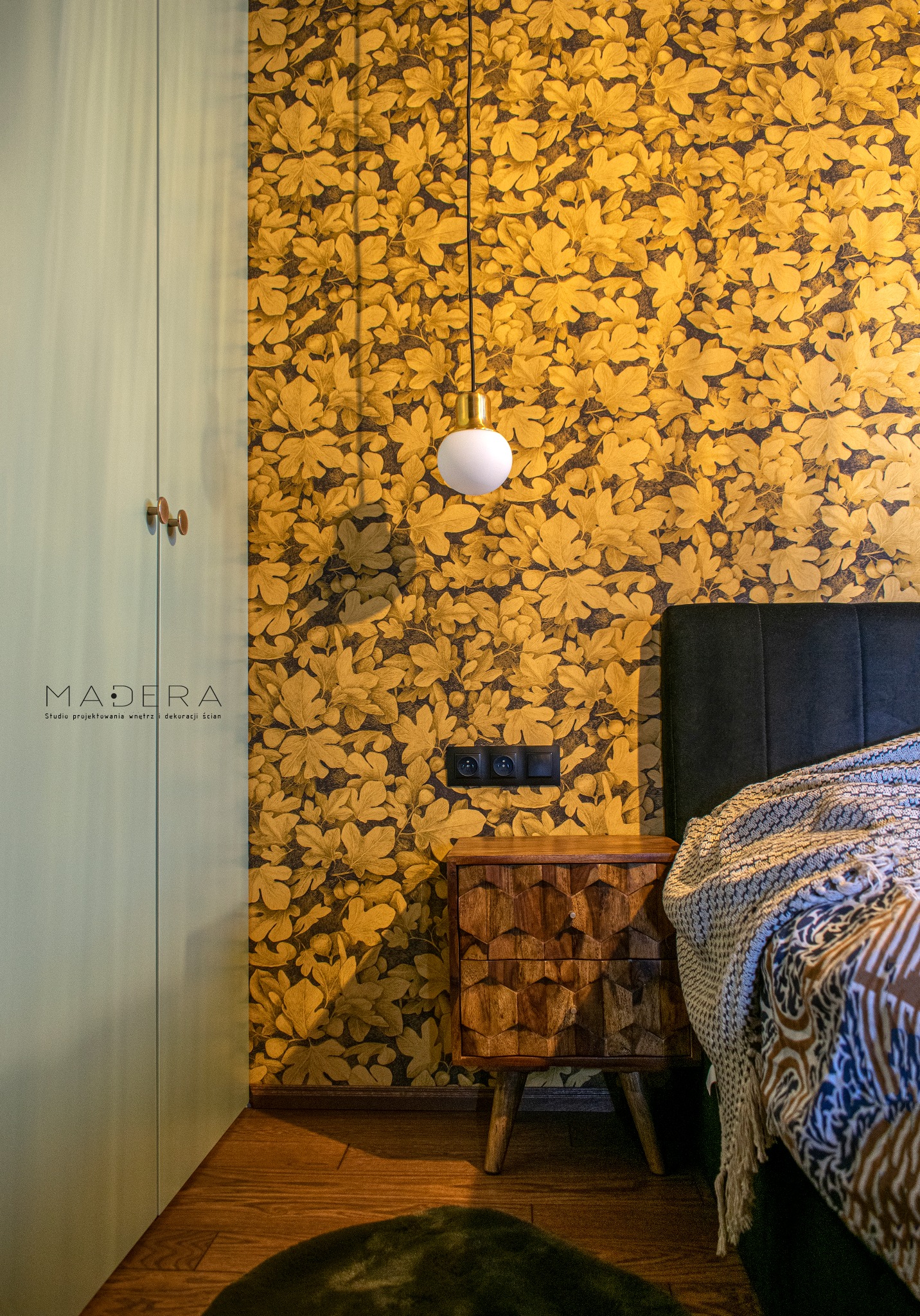 Sypialnia z tapetą w liście, drewnianą szafką nocną i lampą wiszącą. Widoczny fragment łóżka i szafy. Ciepłe oświetlenie, dekoracyjny styl.