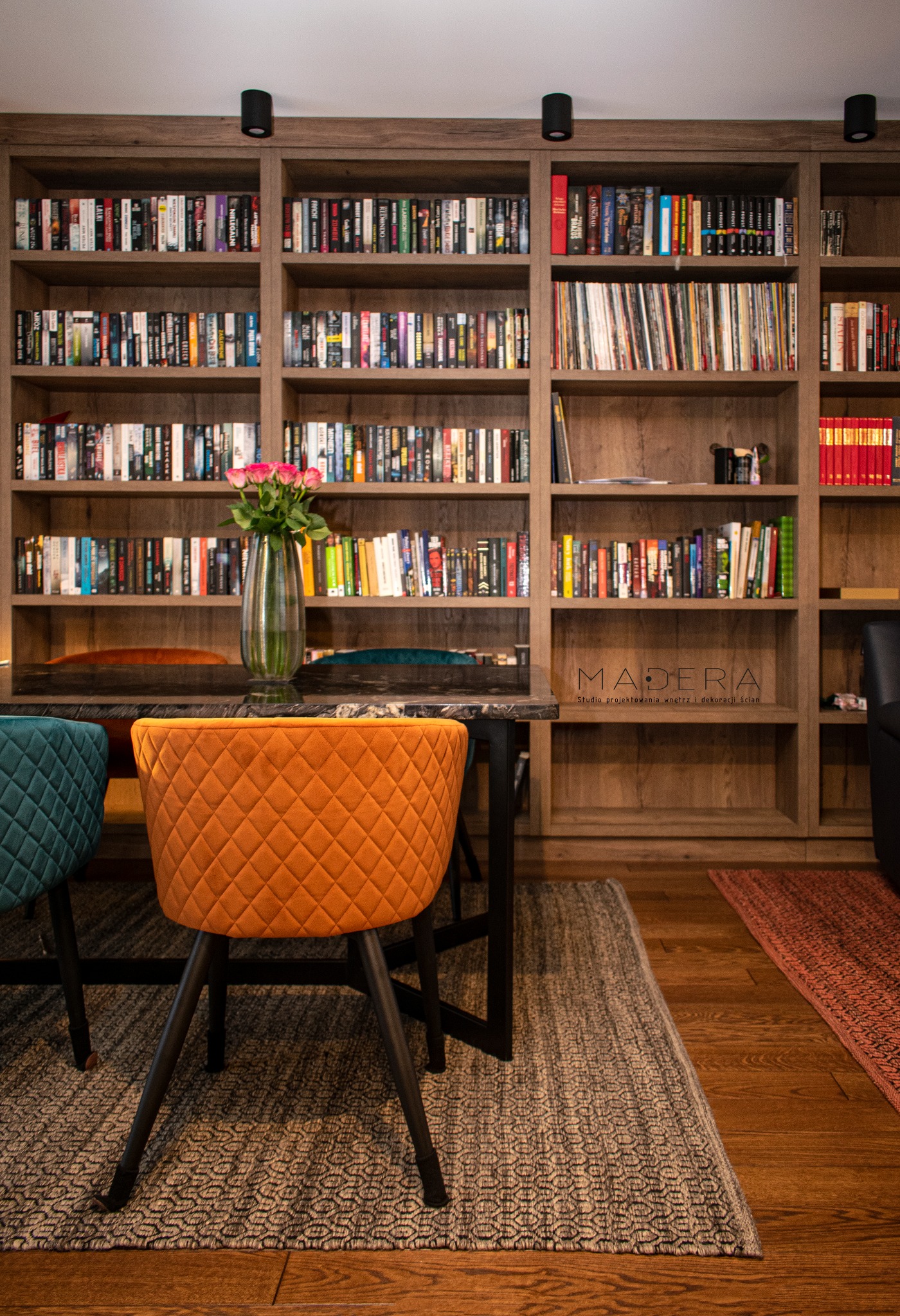 Stylowa aranżacja jadalni w Olsztynie: drewniany regał z książkami, stół z kwiatami i kolorowe krzesła. Projekt Madera, studio aranżacji wnętrz.