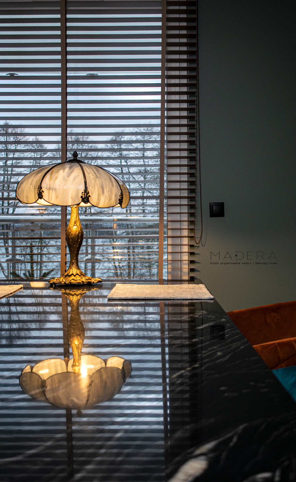 Stylowa lampa na stole z czarnego marmuru, odbijająca się w blacie. W tle okno z drewnianymi żaluzjami i widok na zimowy krajobraz. Logo studia projektowania wnętrz.