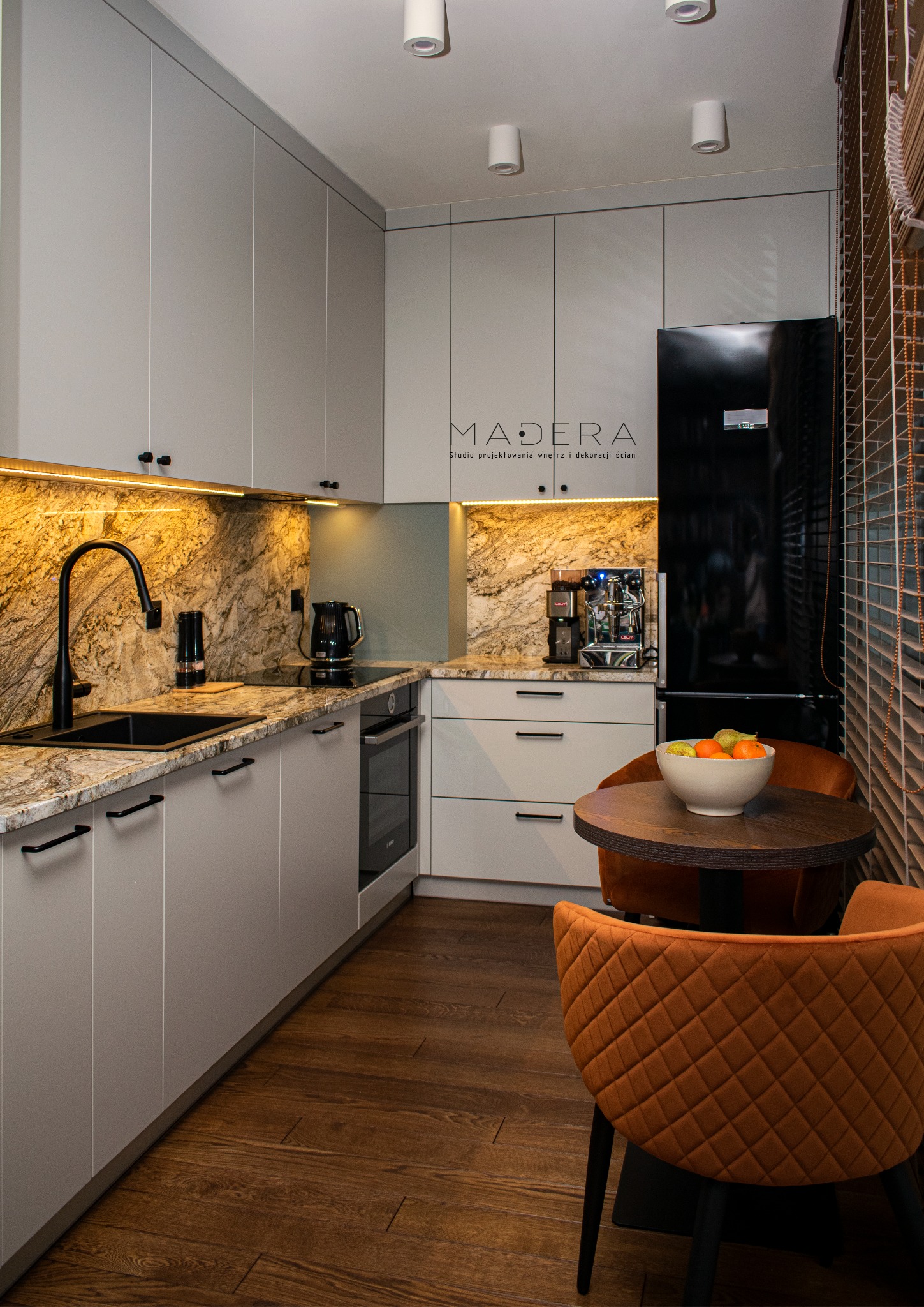 Nowoczesna kuchnia z szarymi szafkami, blatem z marmuru, czarną armaturą i okrągłym stołem z pomarańczowymi krzesłami. Oświetlenie LED podkreśla unikalny charakter wnętrza.