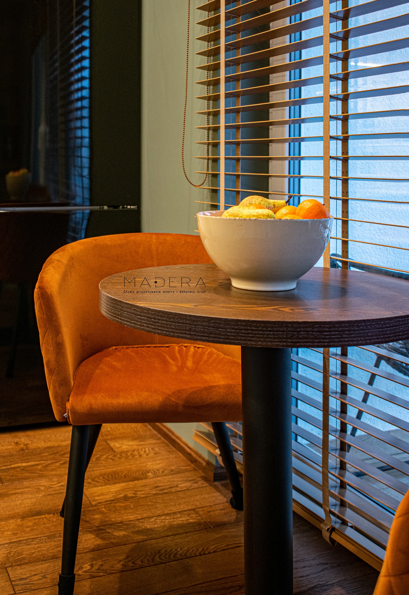 Pomarańczowe krzesło z aksamitu przy okrągłym stoliku z owocami, drewniane żaluzje w tle. Widok na fragment wnętrza, logo firmy projektującej wnętrza na blacie.