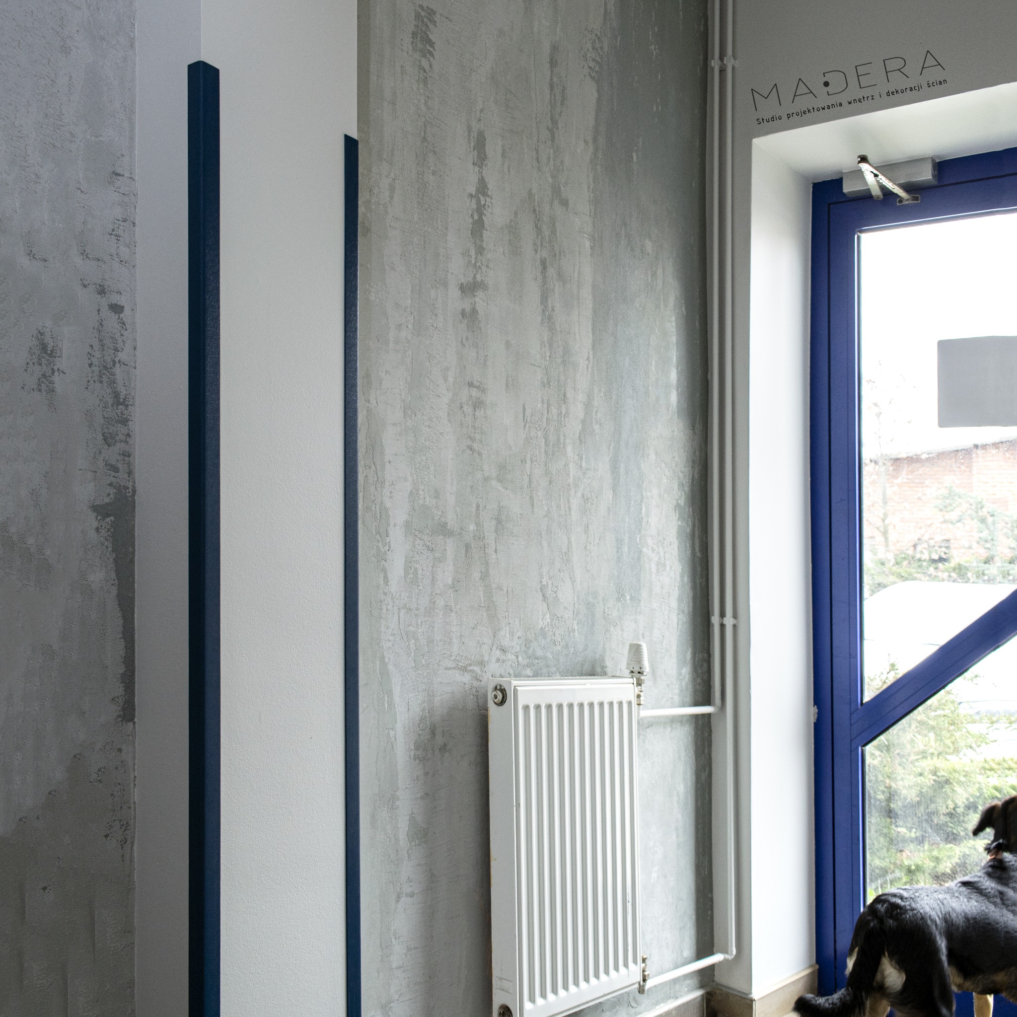 Wnętrze z szarą ścianą o fakturze betonu, biały grzejnik, niebieskie obramowanie drzwi i element dekoracyjny. Pies spogląda przez okno. Logo firmy w prawym górnym rogu.
