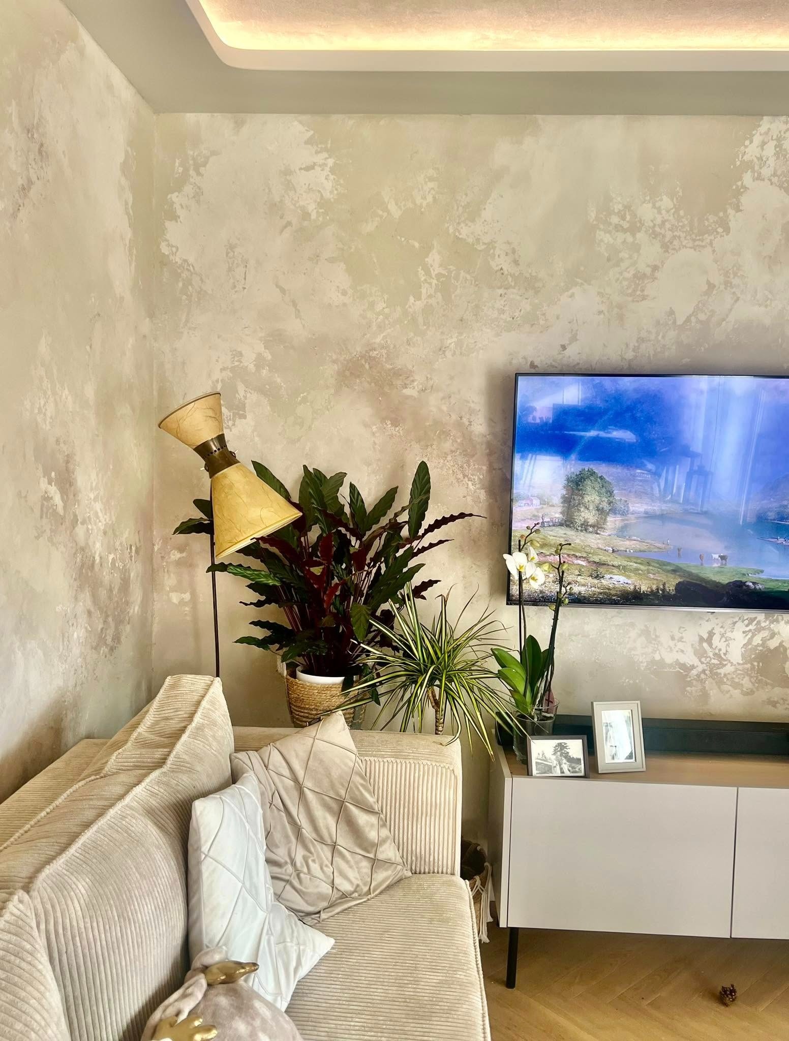 Beżowa sofa z poduszkami, lampa i rośliny na tle ściany z efektem dekoracyjnym w salonie. Telewizor wyświetla krajobraz.