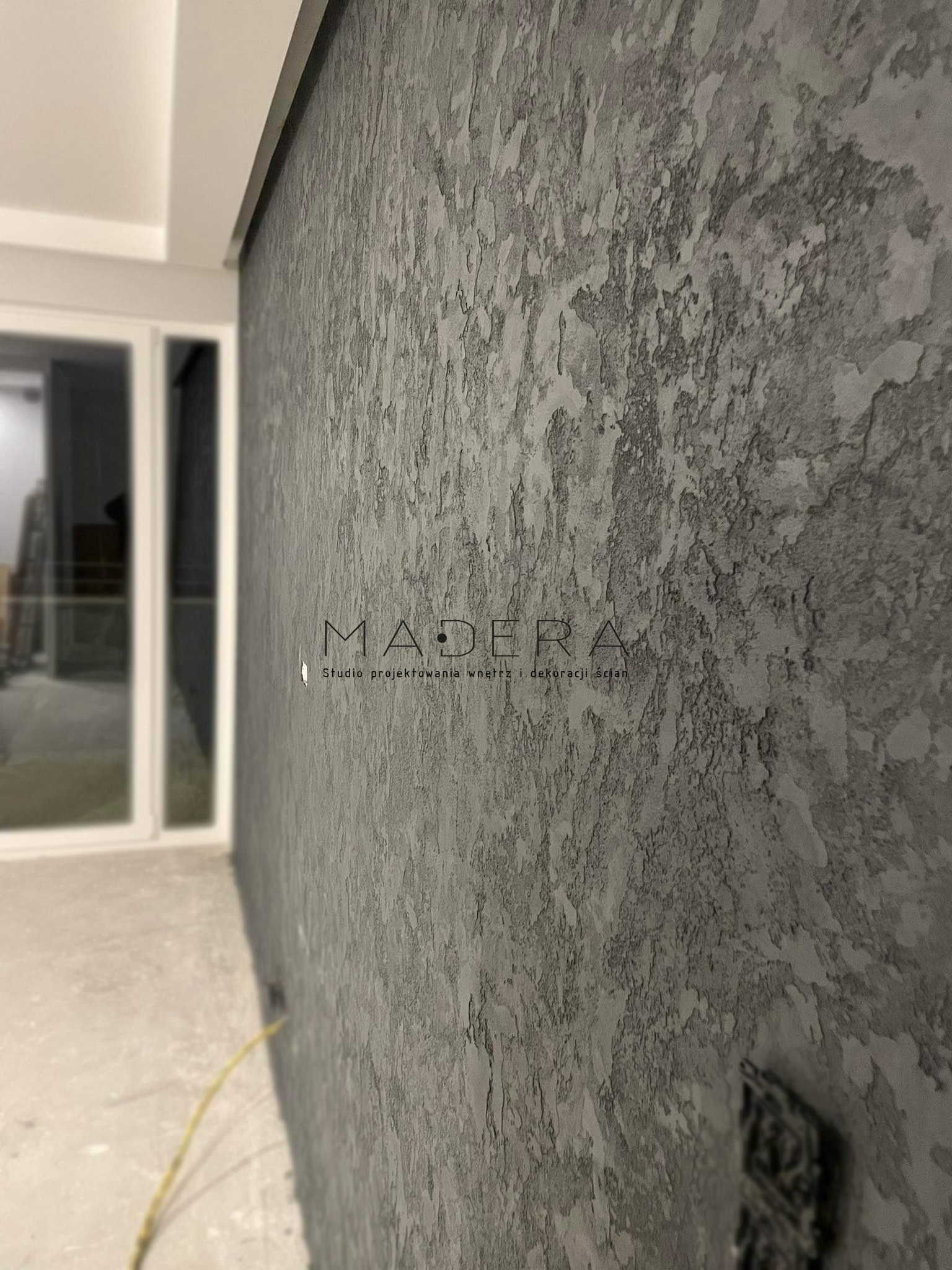 Szara ściana z unikalną fakturą, wykończona w stylu dekoracyjnym. Widoczne logo studia projektowania wnętrz 'MADERA' i fragment okna w tle.
