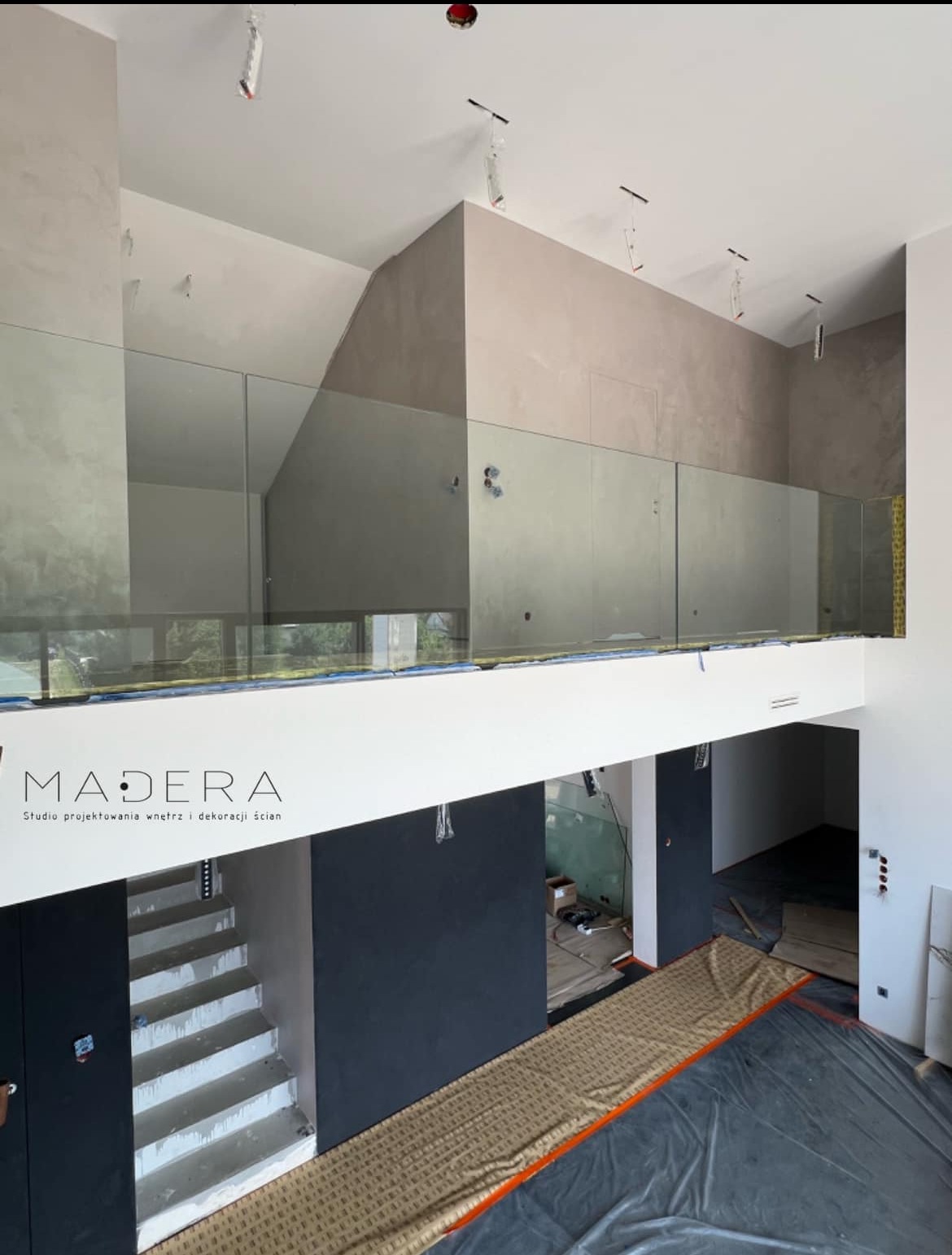 Wnętrze w trakcie wykańczania z antresolą i szklaną balustradą. Widoczne schody, surowe ściany z fakturą i przygotowane podłoże. Logo firmy na pierwszym planie.