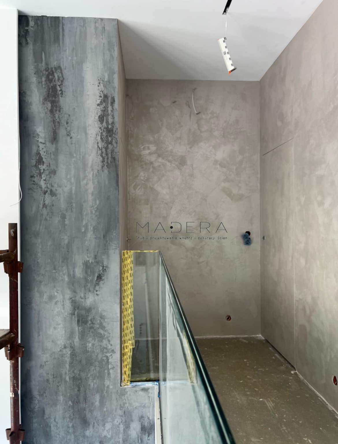 Wnętrze z dekoracyjnymi szarymi ścianami wykończonymi w stylu betonu architektonicznego i szklaną balustradą. Widoczne przygotowania do montażu oświetlenia.