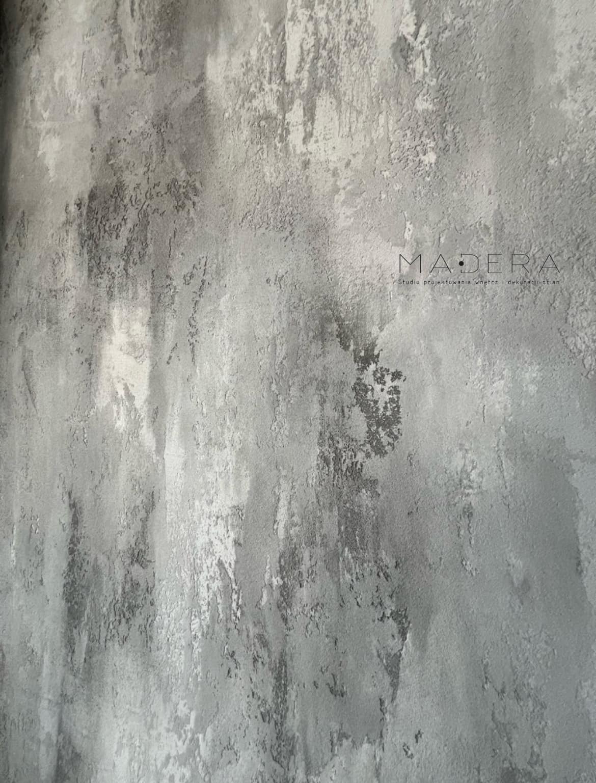 Szara ściana z efektem dekoracyjnym, imitującym przetarcia i nierówności, z subtelnym logo „Madera” w prawym górnym rogu.