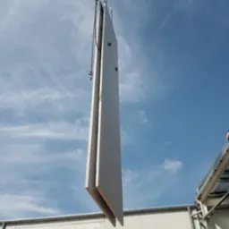 Prefabrykowany element betonowy zawieszony na łańcuchach dźwigu, gotowy do montażu na placu budowy, widoczne niebieskie niebo i fragment konstrukcji budynku.