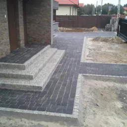 Nowo ułożona nawierzchnia z ciemnej kostki brukowej z jasnymi obrzeżami, prowadząca do schodów wejściowych z jasnego kamienia, widoczne przygotowane podłoże pod dalsze prace brukarskie.