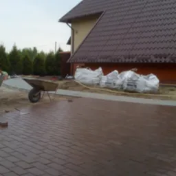 Układanie brązowej kostki brukowej na posesji, widoczna taczka, worki z materiałem budowlanym i narzędzia brukarskie, dom w tle.