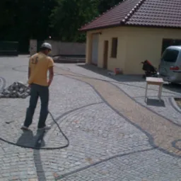 Ukończona nawierzchnia z kostki granitowej ułożona w fantazyjne wzory z jasnym pasem pośrodku, widoczny pracownik w oddali i budynek w tle.