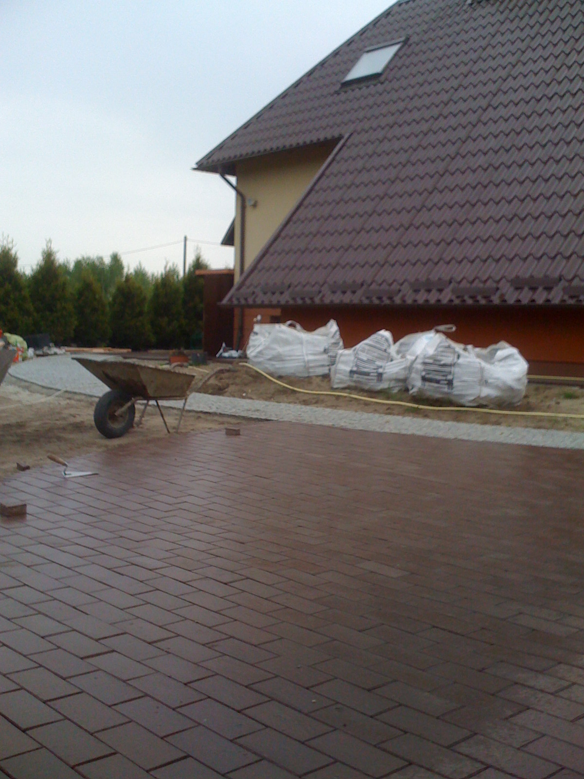 Układanie brązowej kostki brukowej na posesji, widoczna taczka, worki z materiałem budowlanym i narzędzia brukarskie, dom w tle.