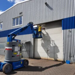 Pracownik w żółtej koszulce na podnośniku koszowym myje elewację magazynu z blachy trapezowej, widoczny niebieski pas elewacji i błękitne niebo z chmurami.