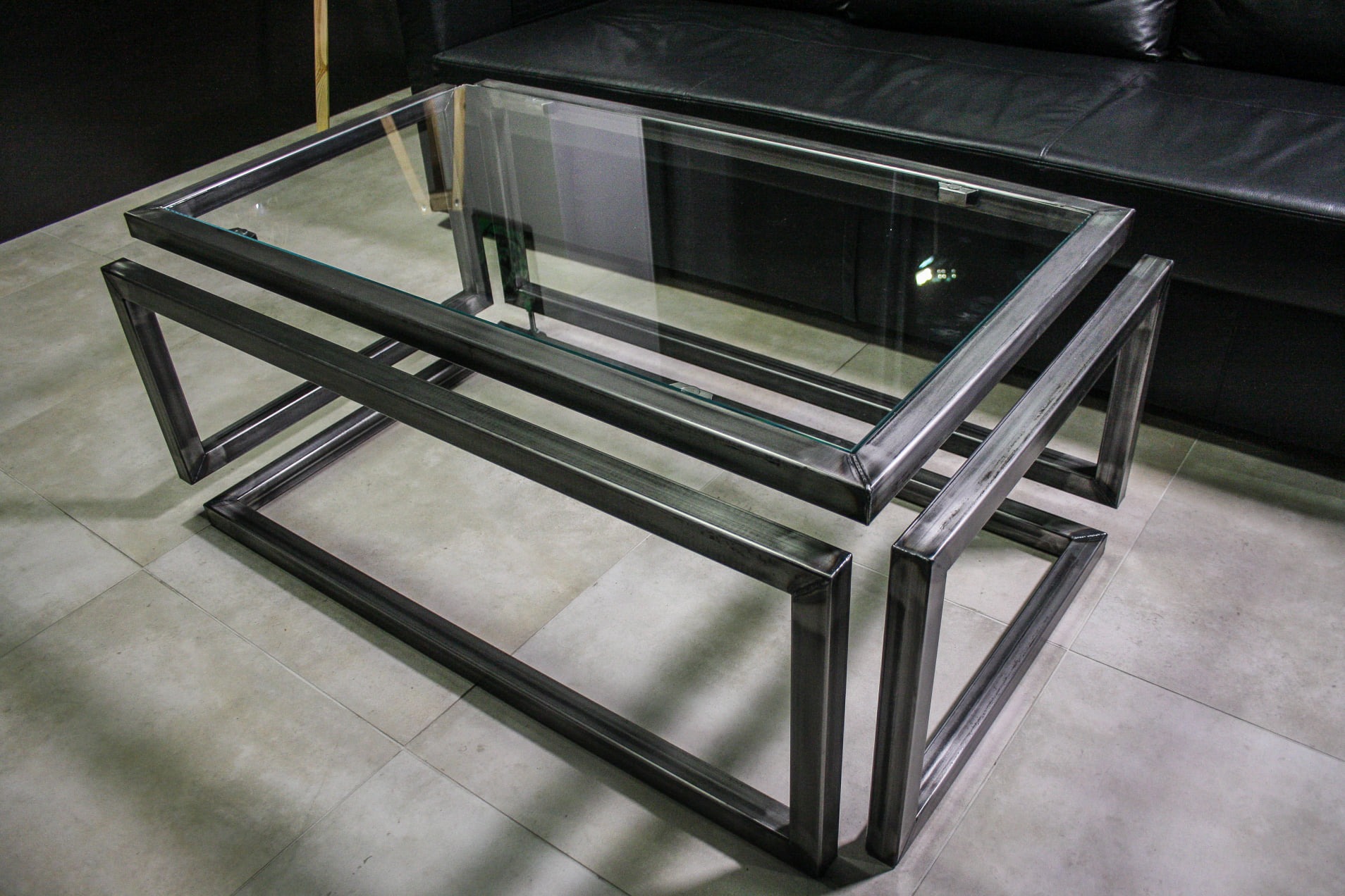 Szklany stolik kawowy z metalową ramą o surowym wykończeniu, wykonany w Gdańsku, na tle czarnej skórzanej sofy.