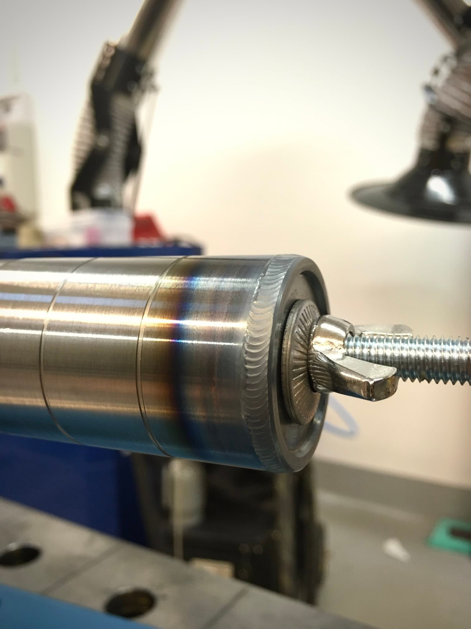 Metalowy cylinder z widocznymi śladami obróbki i tęczowymi przebarwieniami, zamocowany śrubą motylkową w uchwycie, na tle warsztatu.