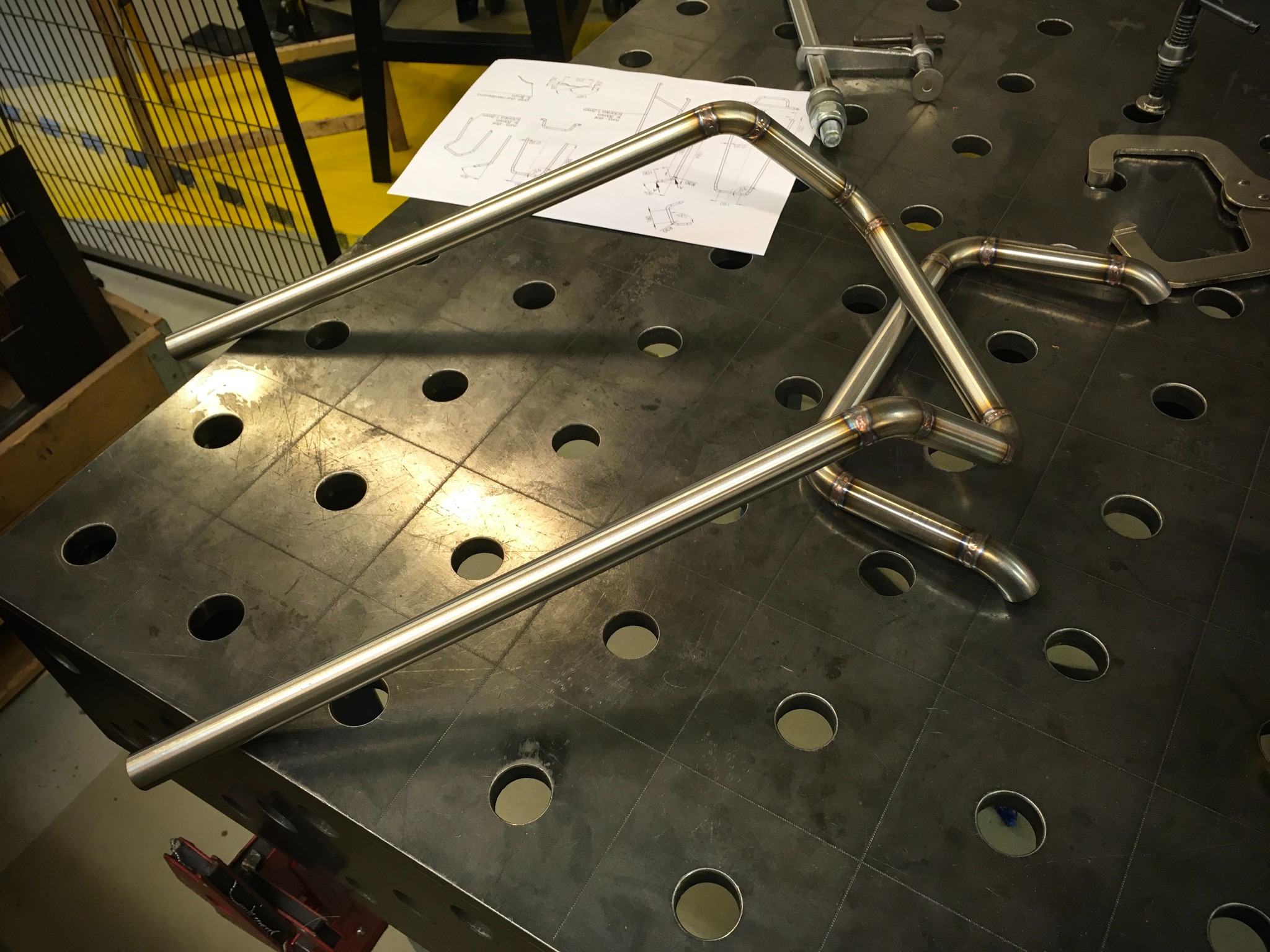 Spawane, gięte rury ze stali nierdzewnej na perforowanym stole spawalniczym, obok schematu i narzędzi.