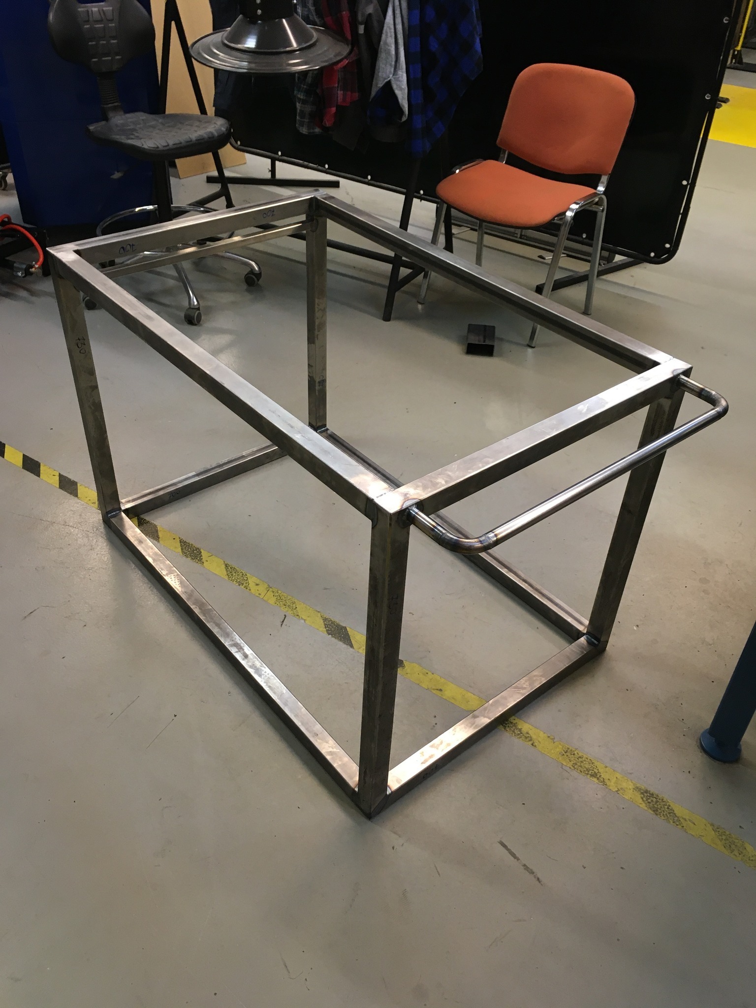 Stalowa rama spawana z profili kwadratowych, z dodatkowym uchwytem z rurki, widoczna na tle szarej podłogi z żółtymi pasami ostrzegawczymi.