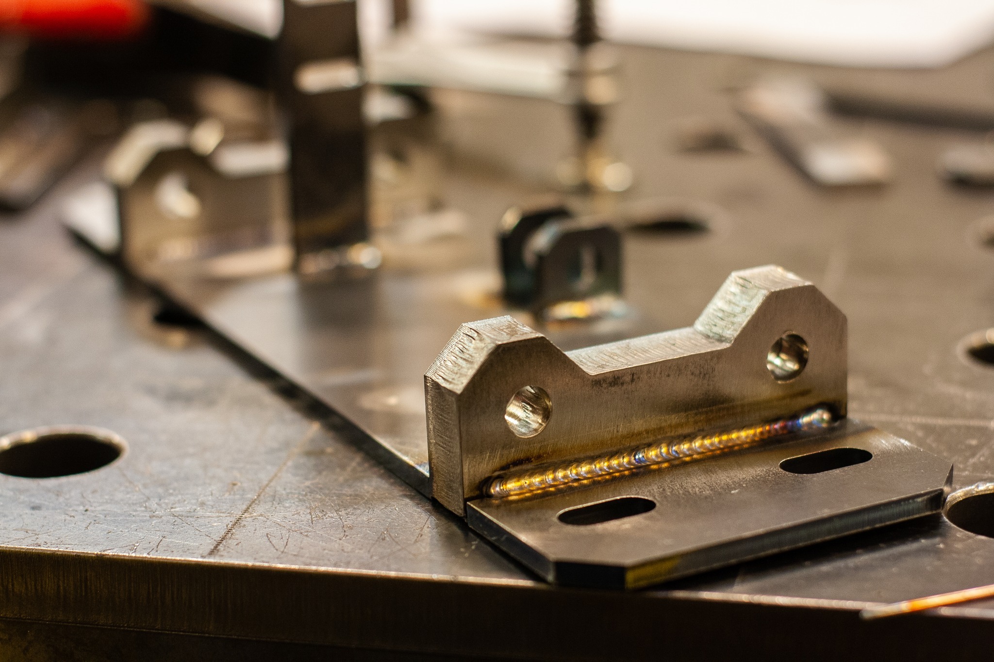 Metalowy element z widocznym spoiwem po procesie łączenia, leżący na metalowym stole z otworami, w tle inne elementy metalowe.