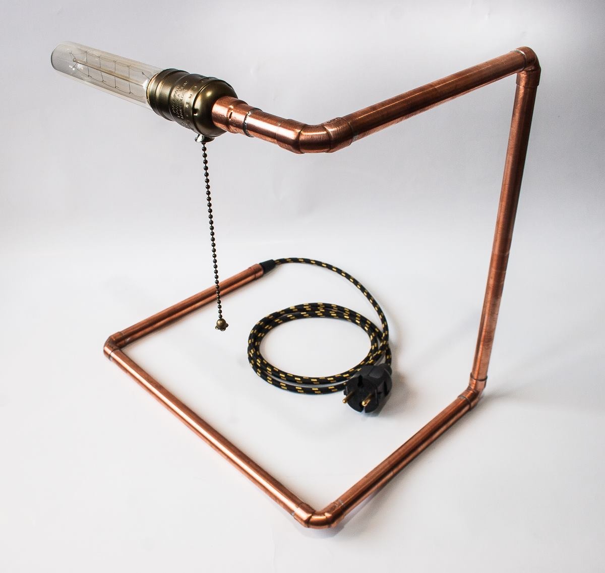 Lampa biurkowa wykonana z miedzianych rur, z żarówką Edison na górze i włącznikiem łańcuszkowym, stojąca na białym tle.