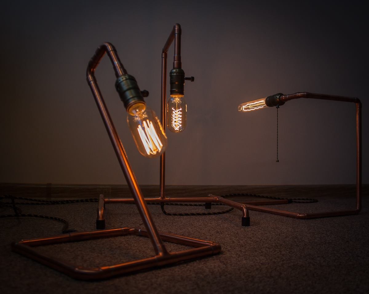 Trzy lampy stołowe w stylu industrialnym, wykonane z miedzianych rurek i żarówek Edisona, ustawione na ciemnym dywanie, z widocznym przewodem w oplocie.
