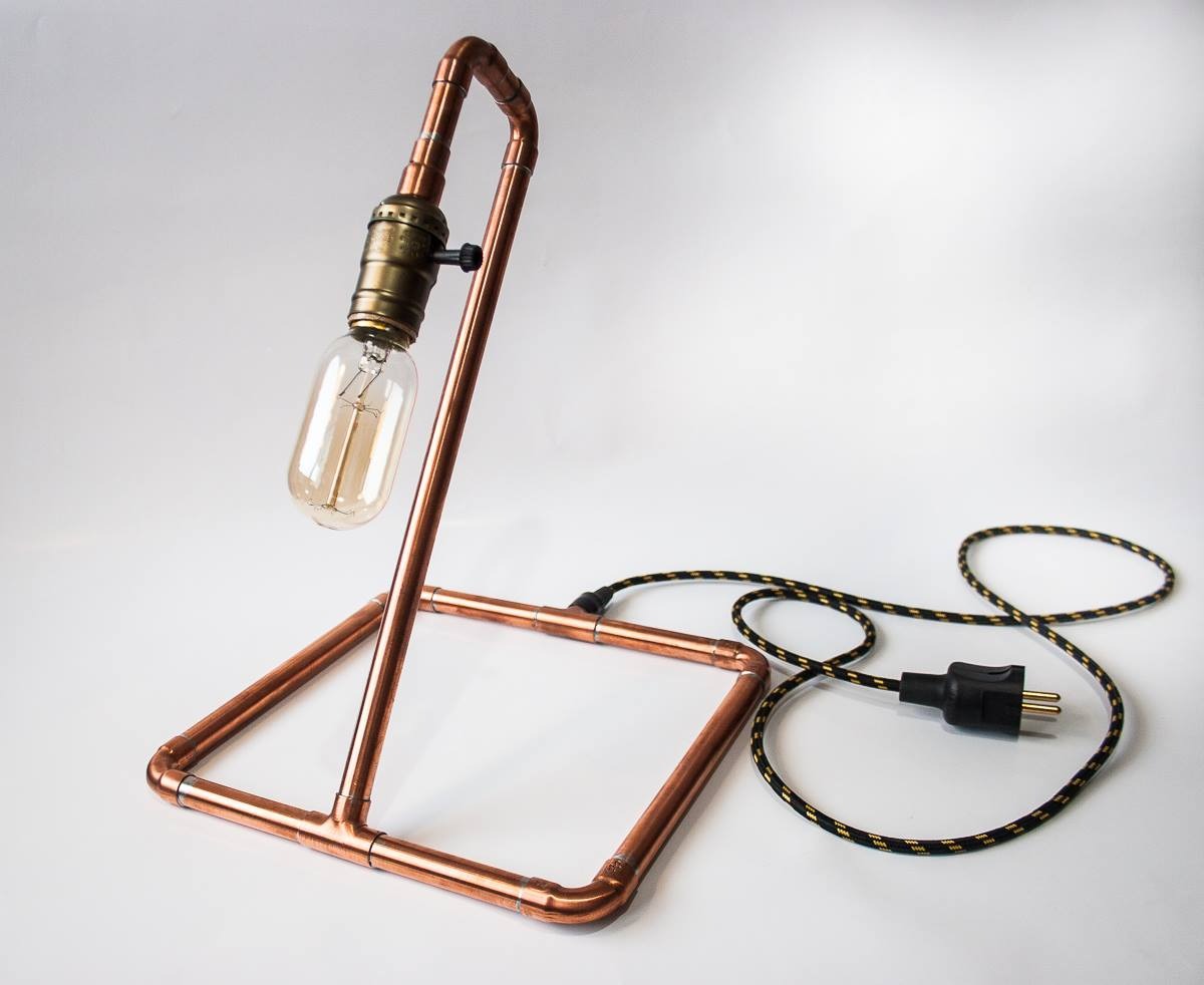 Lampa stołowa wykonana z miedzianych rurek, z żarówką Edison i czarno-złotym kablem w oplocie, na białym tle.
