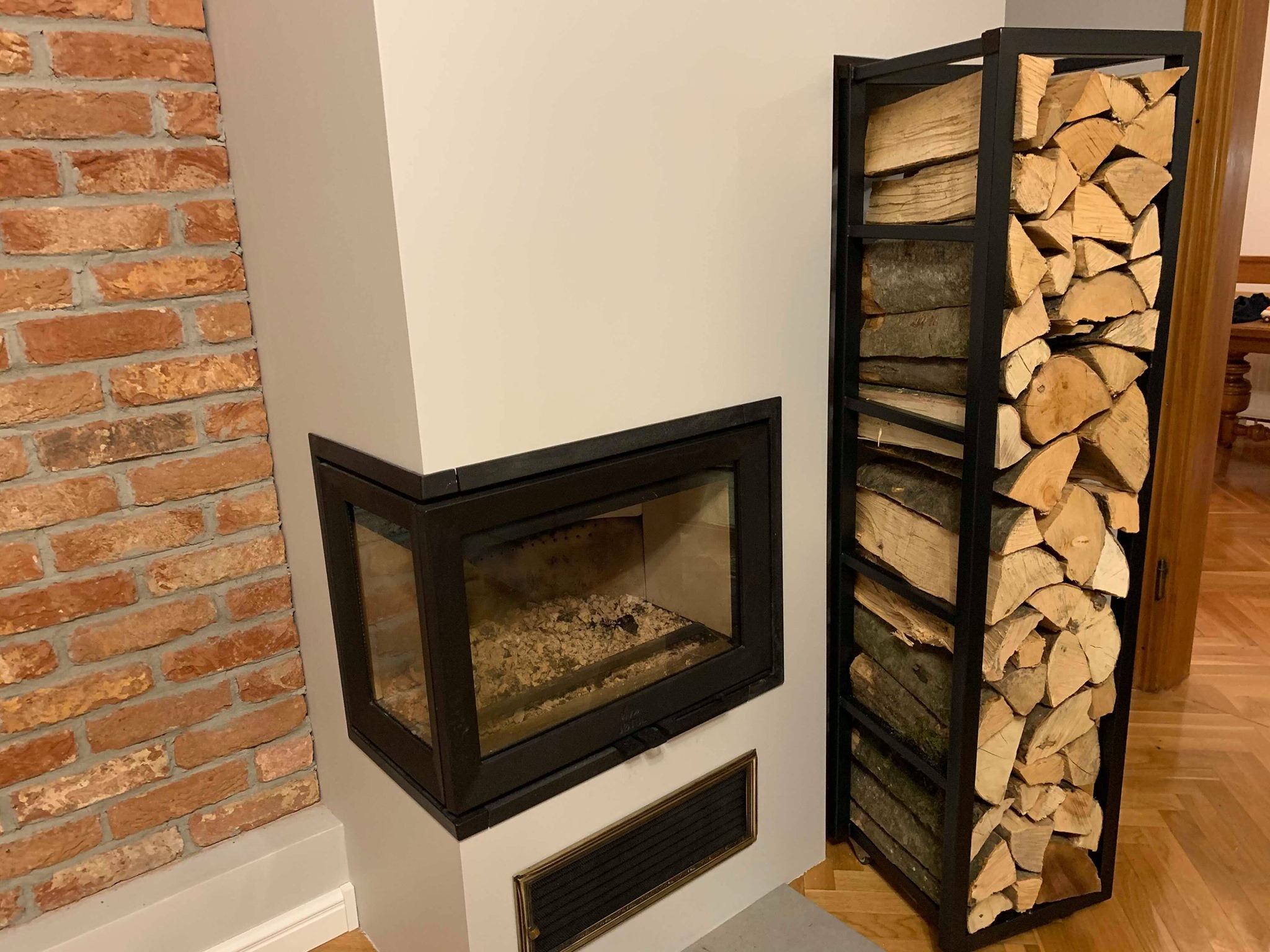Nowoczesny kominek narożny z czarną obudową, obok metalowy regał z ułożonym drewnem opałowym, ściana z cegły i parkiet na podłodze.