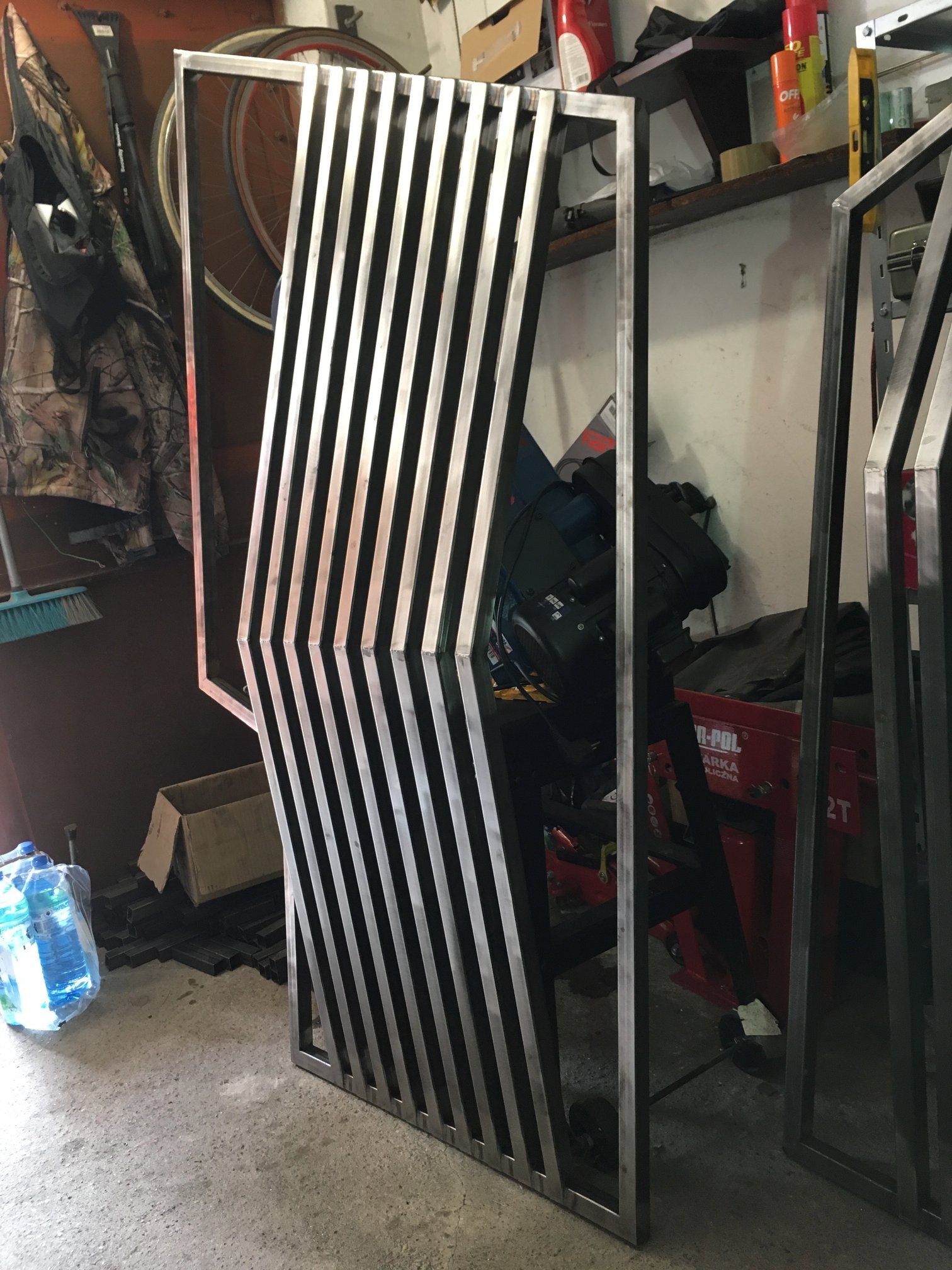 Metalowa konstrukcja z pionowymi prętami, spawana rama, surowe wykończenie, widoczne ślady obróbki, tło warsztatowe z narzędziami i akcesoriami.