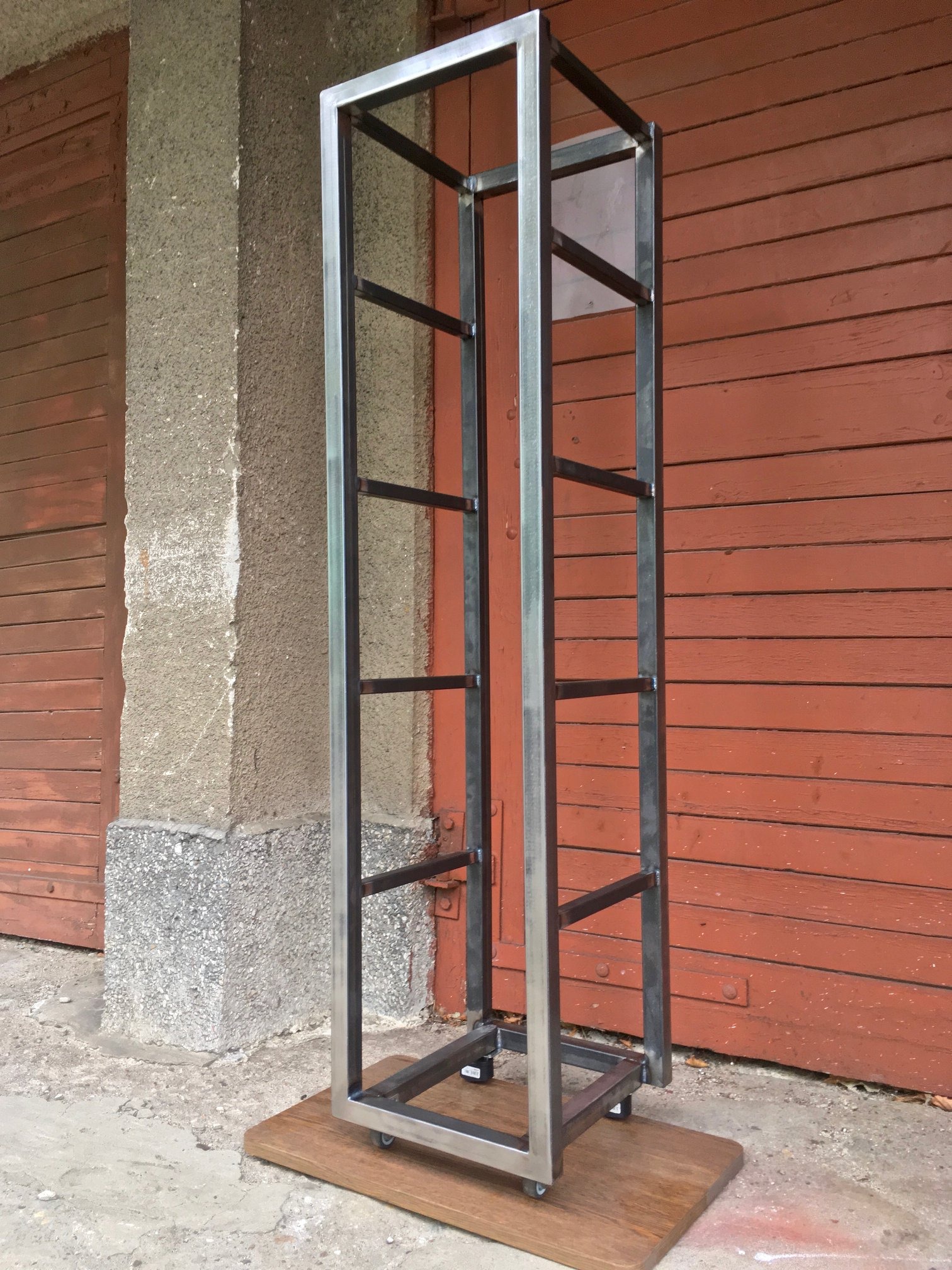 Spawana metalowa konstrukcja regału na kółkach, surowy stan, tło to ceglany budynek i betonowy filar.