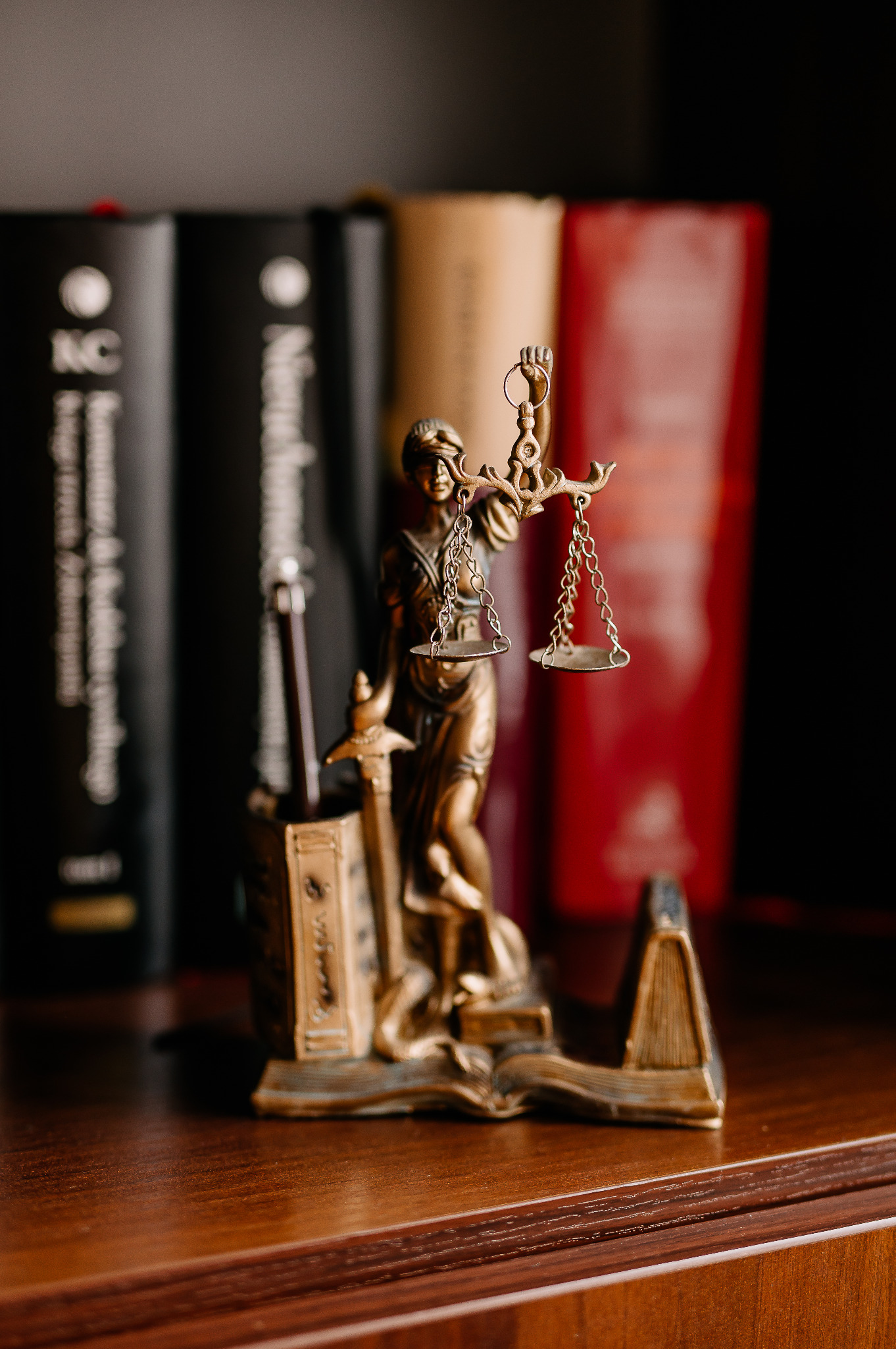 Brązowa figurka Temidy z wagą i mieczem na tle książek na drewnianym biurku.