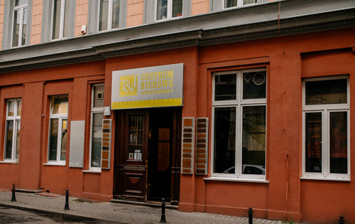 Elewacja budynku biurowego z szyldem 'RJ Centrum Biurowe' nad wejściem, widoczne okna z białymi ramami i ciemne drewniane drzwi wejściowe.