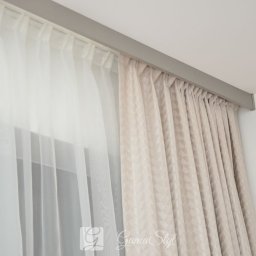 Dekoracje okien | Karnisze, Rolety, Żaluzje | GAMA STYL - Dwie warstwy zasłon: przejrzysta, biała woalowa i beżowa z geometrycznym wzorem, zawieszone na szynie podsufitowej w minimalistycznym wnętrzu.