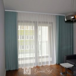 Dekoracje okien | Karnisze, Rolety, Żaluzje | GAMA STYL - Eleganckie, turkusowe zasłony panelowe z białymi, zwiewnymi firanami w salonie w Warszawie, z widoczną nowoczesną lampą wiszącą i fragmentem szarej sofy.