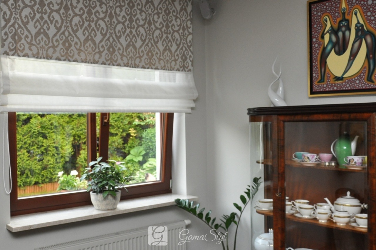 Elegancka roleta rzymska w odcieniach bieli i beżu z dekoracyjnym wzorem, zamontowana w oknie z widokiem na zielony ogród, uzupełniona doniczką z rośliną na marmurowym parapecie, obok antyczny...