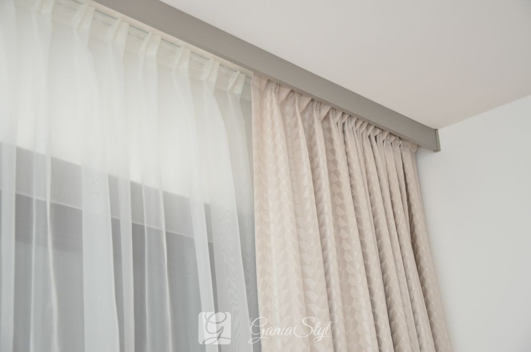 Dwie warstwy zasłon: przejrzysta, biała woalowa i beżowa z geometrycznym wzorem, zawieszone na szynie podsufitowej w minimalistycznym wnętrzu.