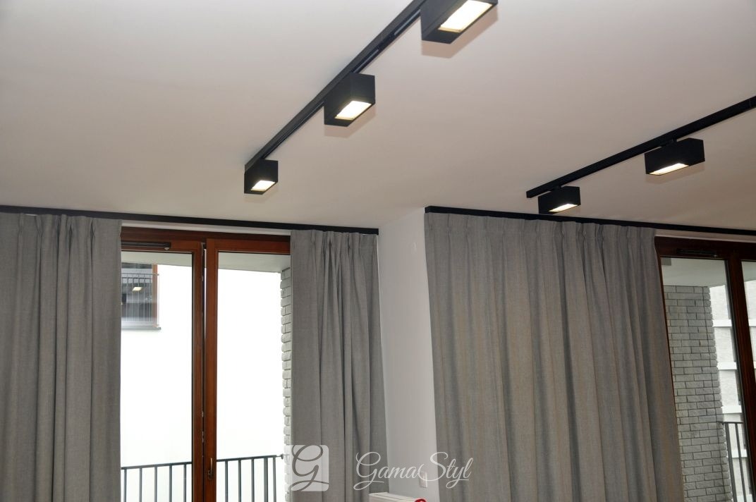 Szare zasłony zaciemniające zawieszone na czarnym karniszu, z widocznymi lampami sufitowymi na szynoprzewodzie.