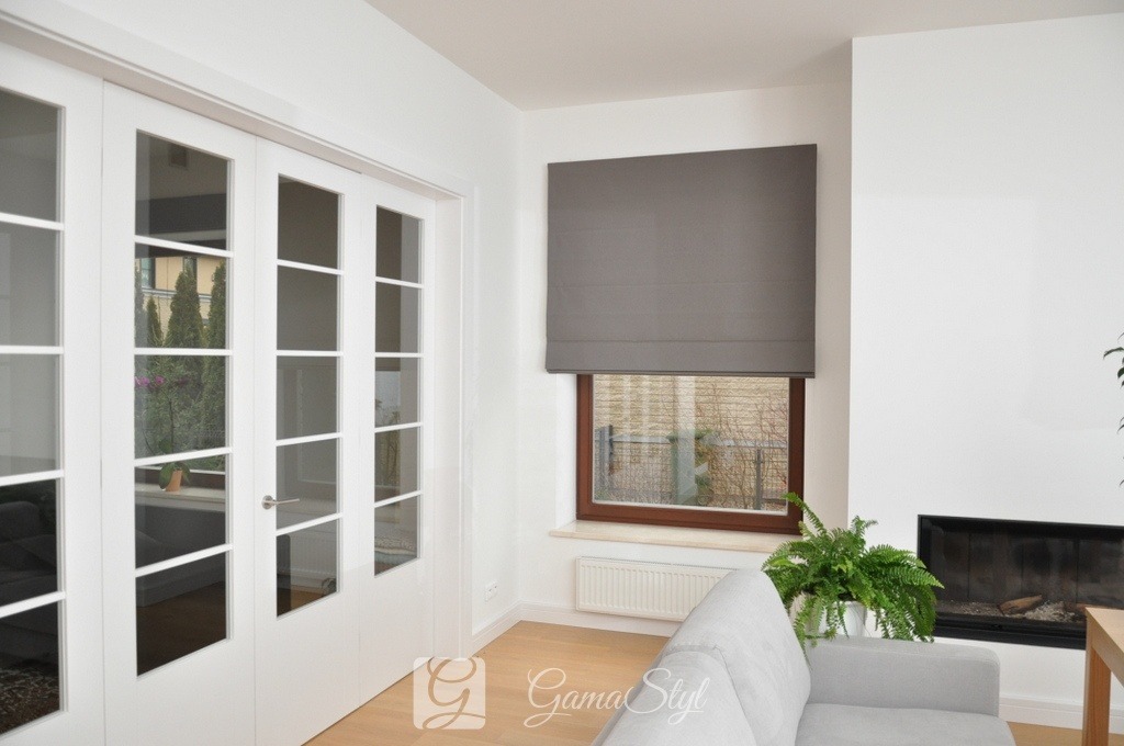 Szara tkanina na oknie, ułożona w poziome fałdy, tworzy dekoracyjny element zaciemniający, w tle białe ściany, przeszklone drzwi i fragment kominka.