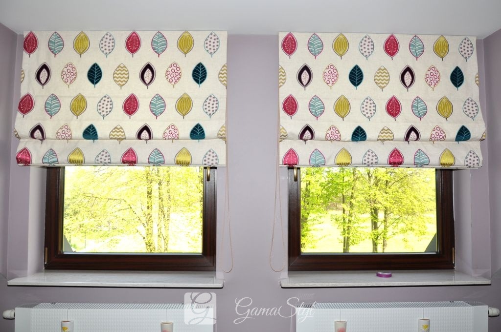 Dwa okna z brązowymi ramami, przesłonięte roletami w stylu rzymskim z motywem kolorowych liści na jasnym tle, widok na zielone drzewa za oknem.